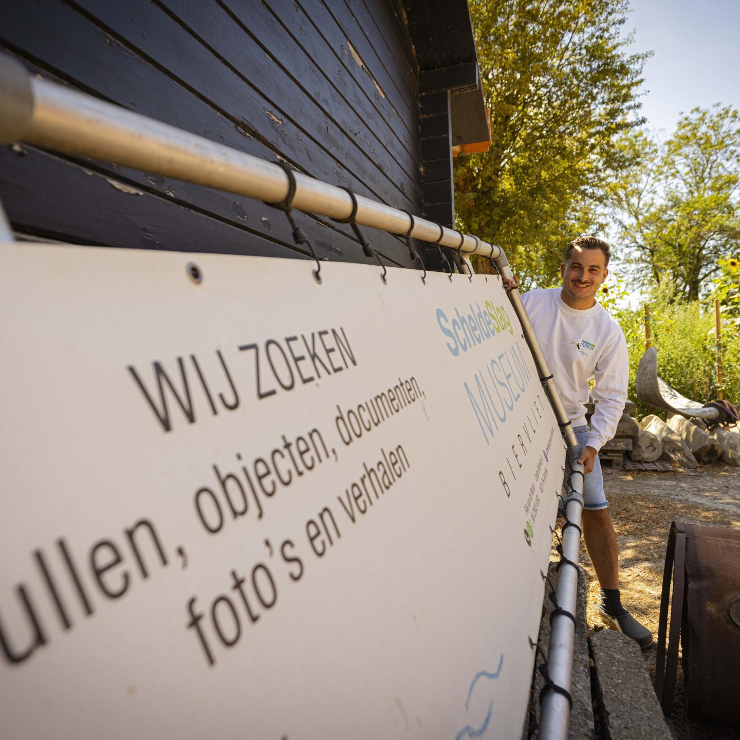 Rik staat bij een aankondigingsbord van ScheldeSlag Museum op het erf in Biervliet. Op het bord staat: Wij zoeken spullen, objecten, documenten, foto's en verhalen.