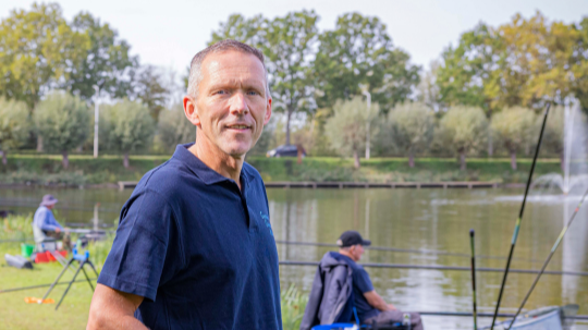 Frank staat op de voorgrond van de visvijver in Axel met vissers op de achtergrond.