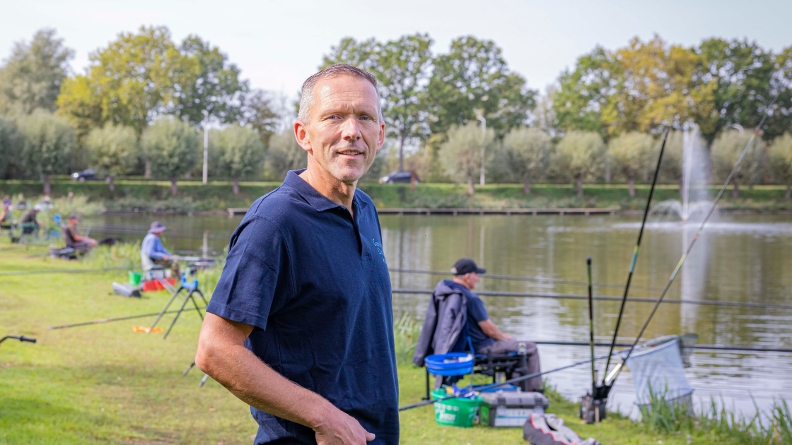 Frank staat op de voorgrond van de visvijver in Axel met vissers op de achtergrond.