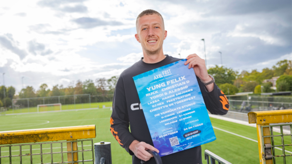 Menno voor een voetbalveld met een poster van Axelfest in zijn hand