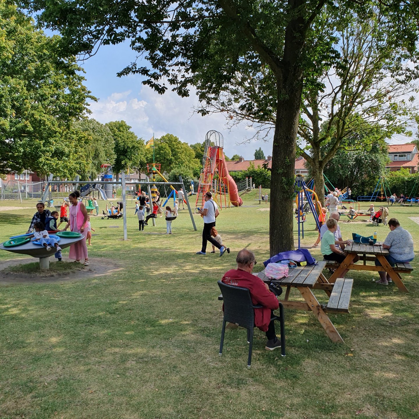Deel van Speeltuin Oranjekwartier: een grasveld met speeltoestellen waar kinderen op spelen en op de voorgrond, onder de bomen, picknicktafels waar volwassenen aan zitten.