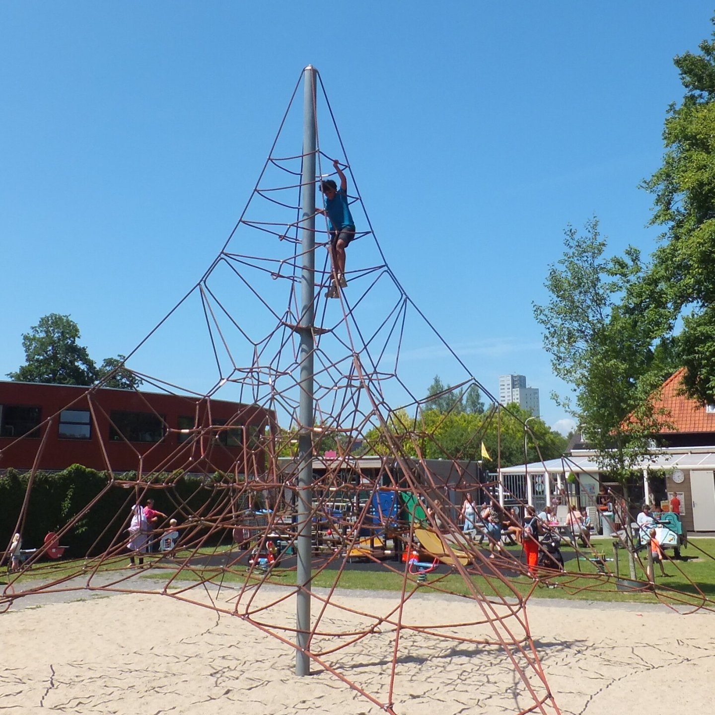 Groot klimtoestel: een piramidevorm van touwen met bovenin een jongetje dat naar de top klimt. Op de achtergrond een terras waar mensen van een drankje en de zon genieten.