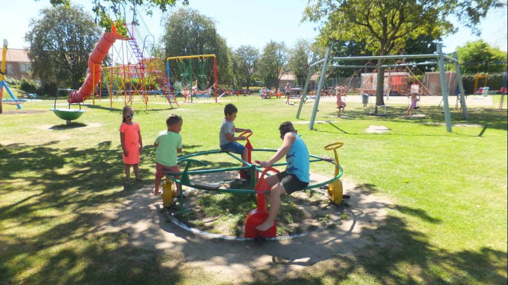 Deel van Speeltuin Oranjekwartier met op de voorgrond een ronddraaiend speeltoestel waar drie kinderen op spelen terwijl een meisje met gesminckt gezicht toekijkt. Op de achtergrond nog andere, kleurrijke speeltoestellen en volwassen bomen.