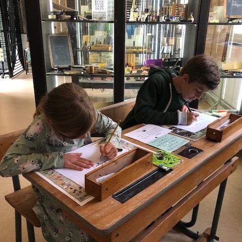 Een meisje en jongen zitten aan een oud schooltafeltje met inktpotje te schrijven in het Schooltijd museum