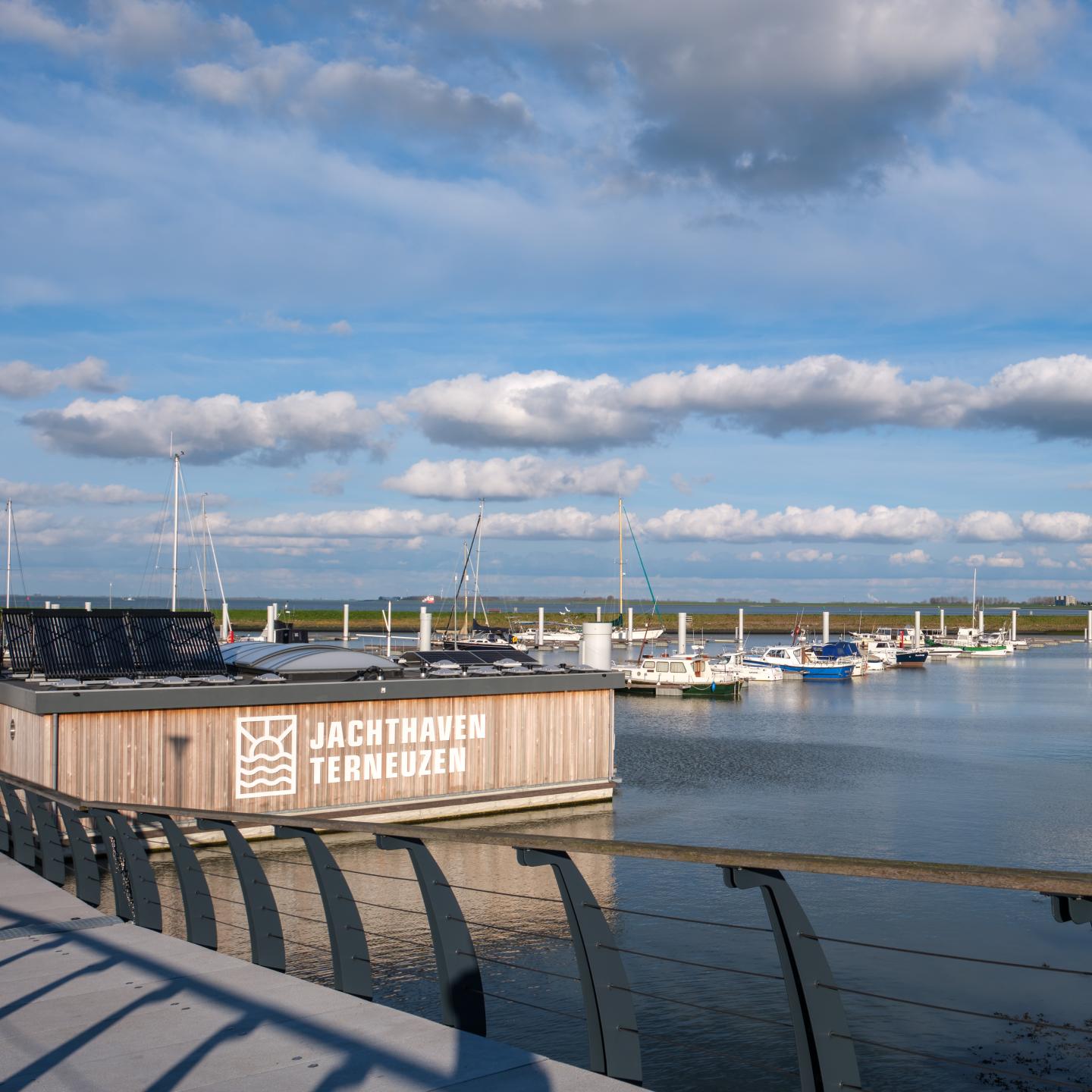 Jachthaven met op voorgrond gebouw met voorzieningen, daarachter schepen aan de steiger en rechts de wal met in de verte een groot appartementengebouw