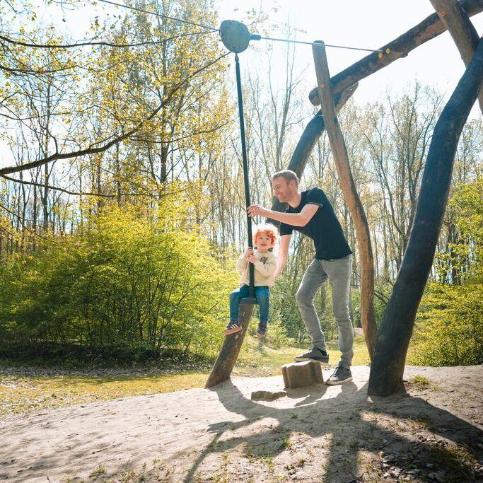 vader helpt jong kindje bij een tokkelbaan in het natuurpark