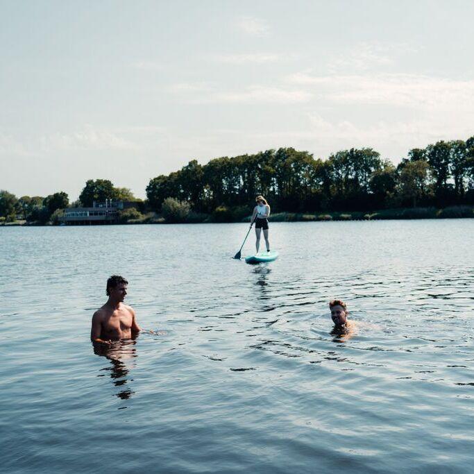 Twee mannen staan en zwemmen in het water met op de achtergrond een meisje op supboard