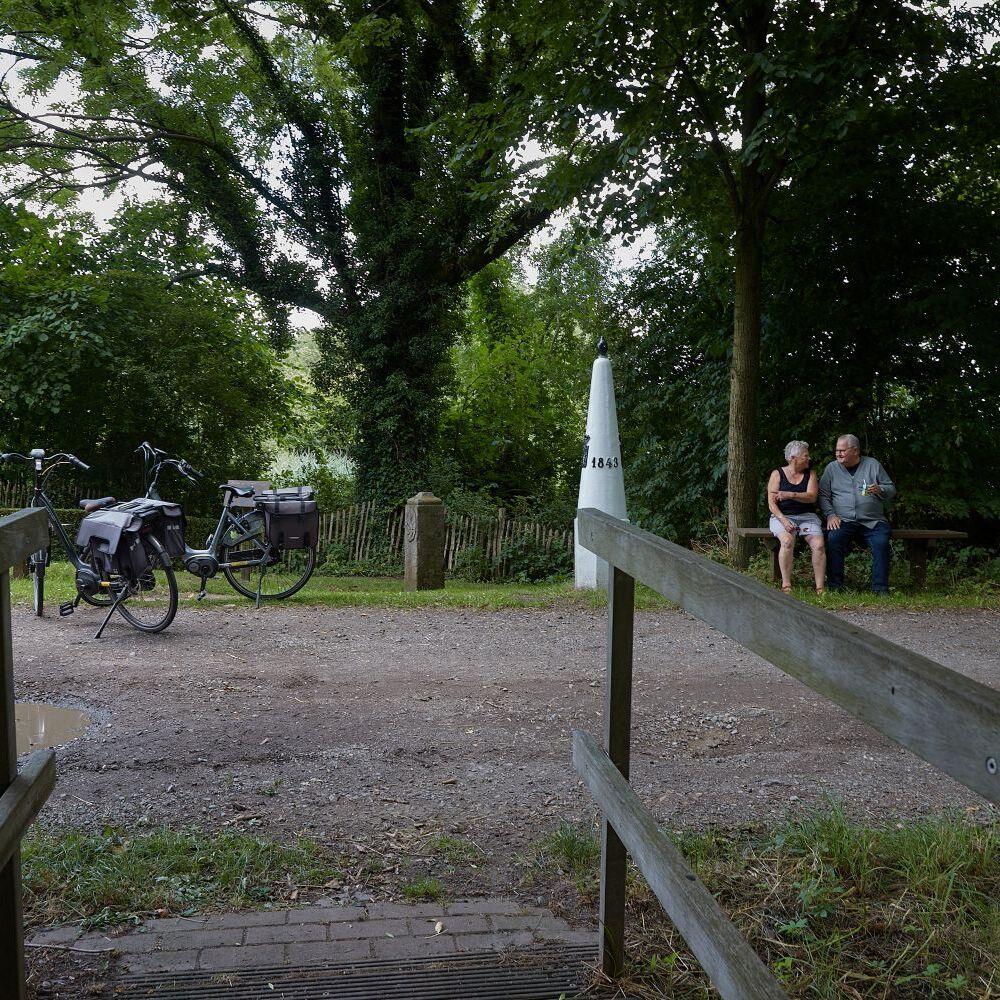 Tweeling grenspalen bij het dorp Overslag met rechts in beeld twee fietsers die samen op een bankje uitrusten