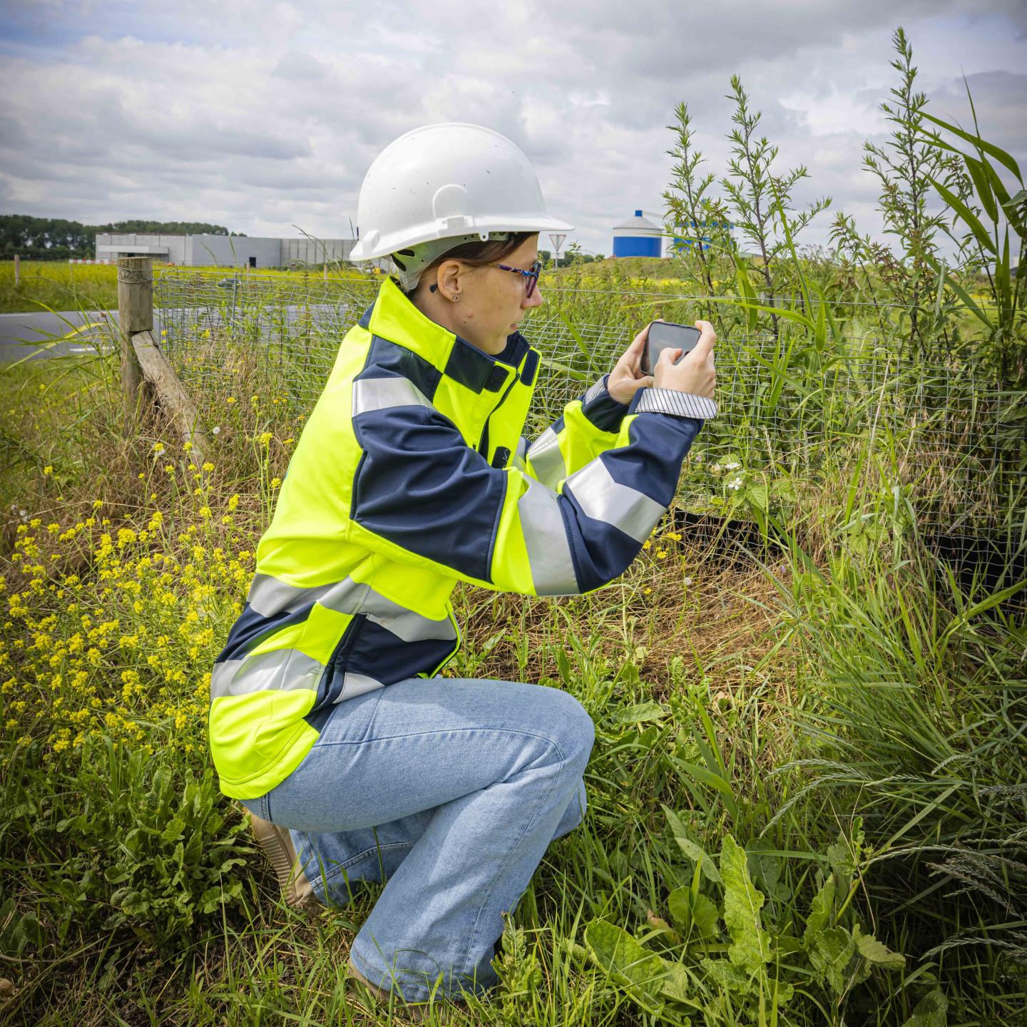 Lieke met veiligheidshelm op en veiligheidskleding aan gehurkt in het veld met mobiel een foto nemend