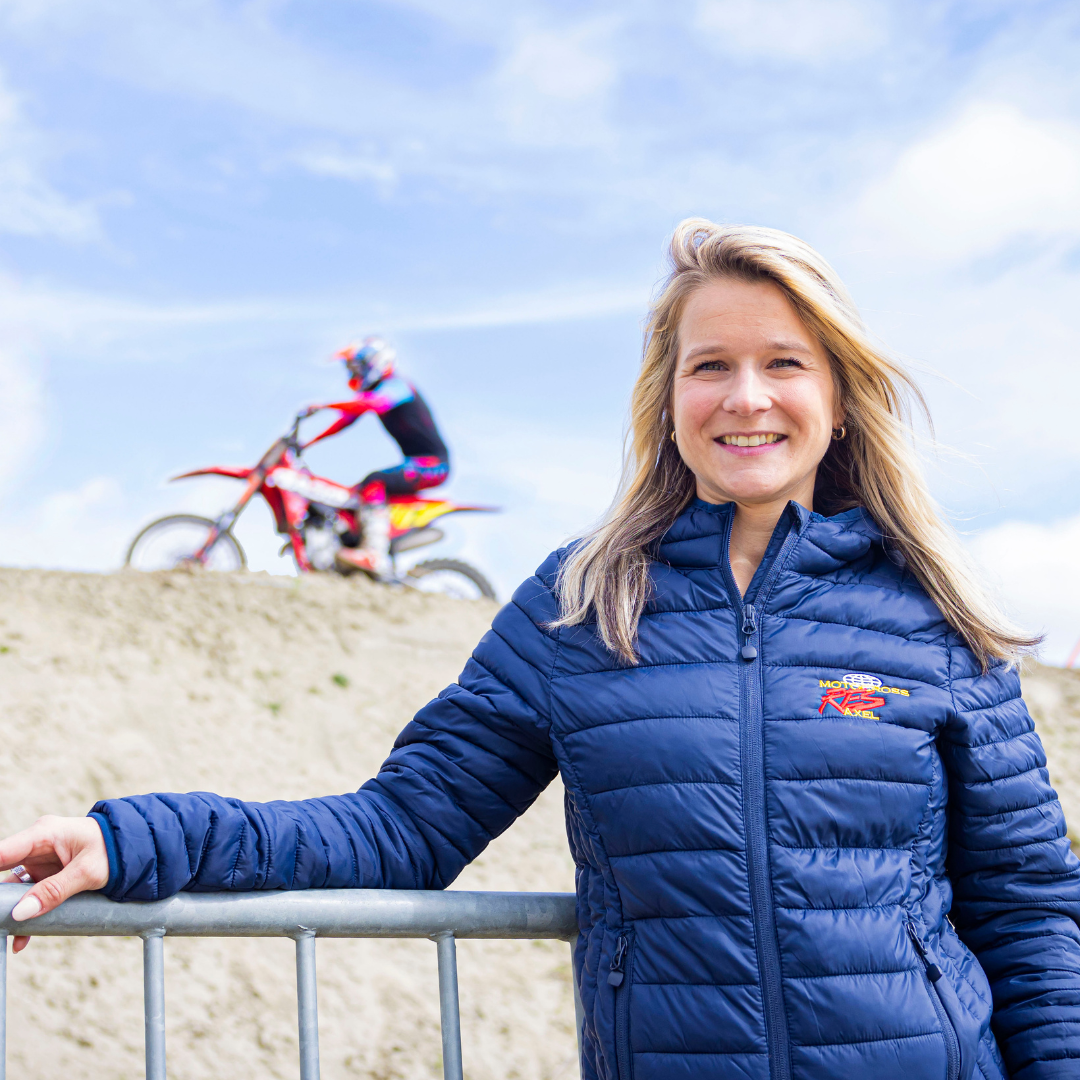 Evie leunt tegen een hek aan en kijkt lachend in de camera terwijl achter haar op een zandberg een motorcrosser voorbij rijdt
