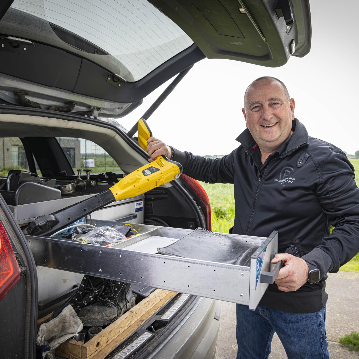 Martin staat onder de achterklep van zijn auto en haalt een stuk gereedschap eruit.