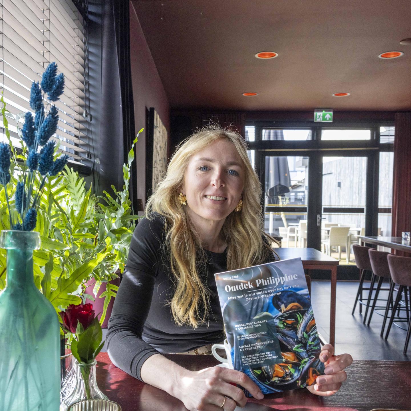 Michelle met een brochure 'Ontdek Philippine' in haar handen poseert voor de camera terwijl ze aan een horecatafeltje zit