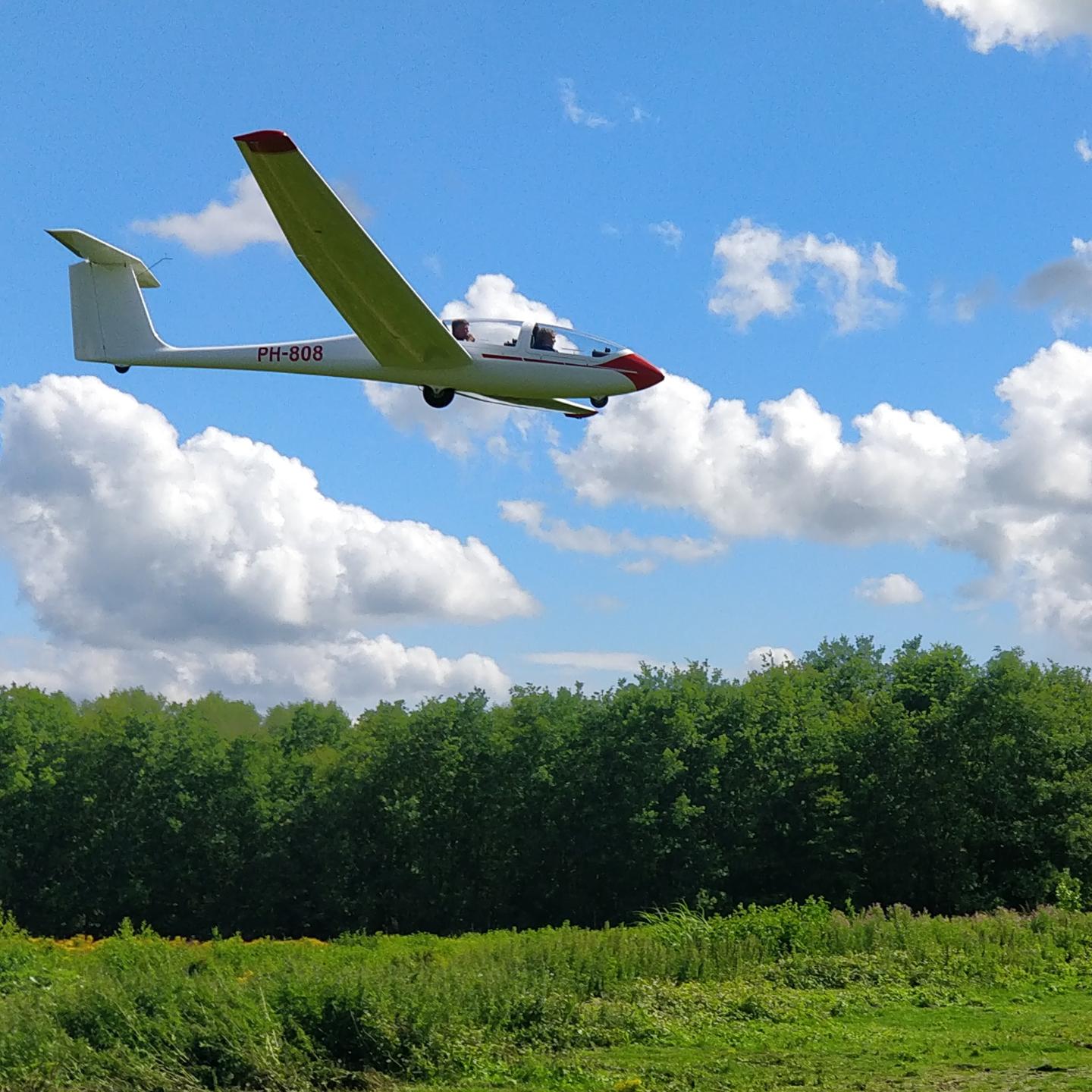 Zweefvliegtuig bezig met landing boven groen veld met groene bomen op de achtergrond