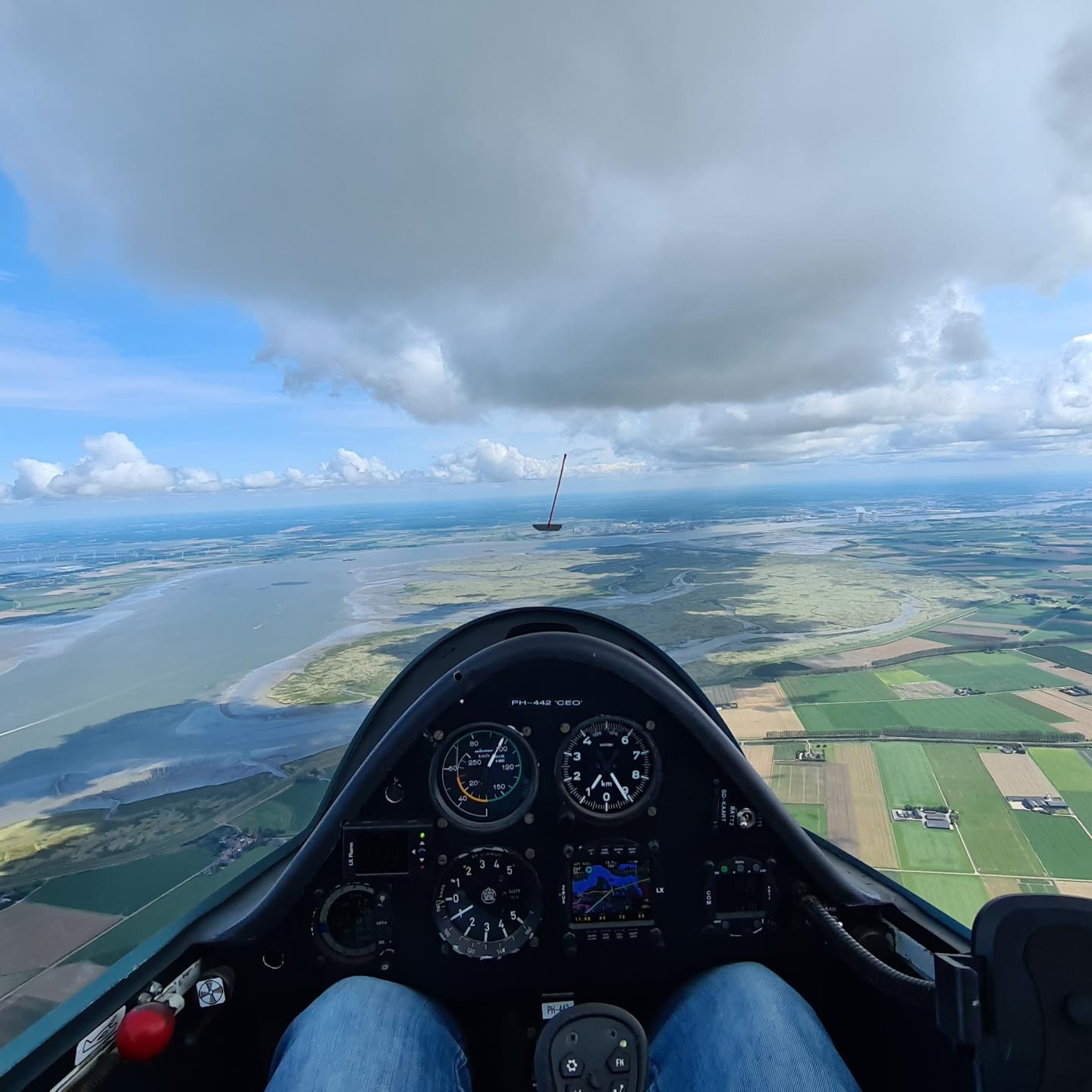 Foto van de cockpit van een zweefvliegtuig met daarboven uit weidse uitzicht hoog boven in de lucht.