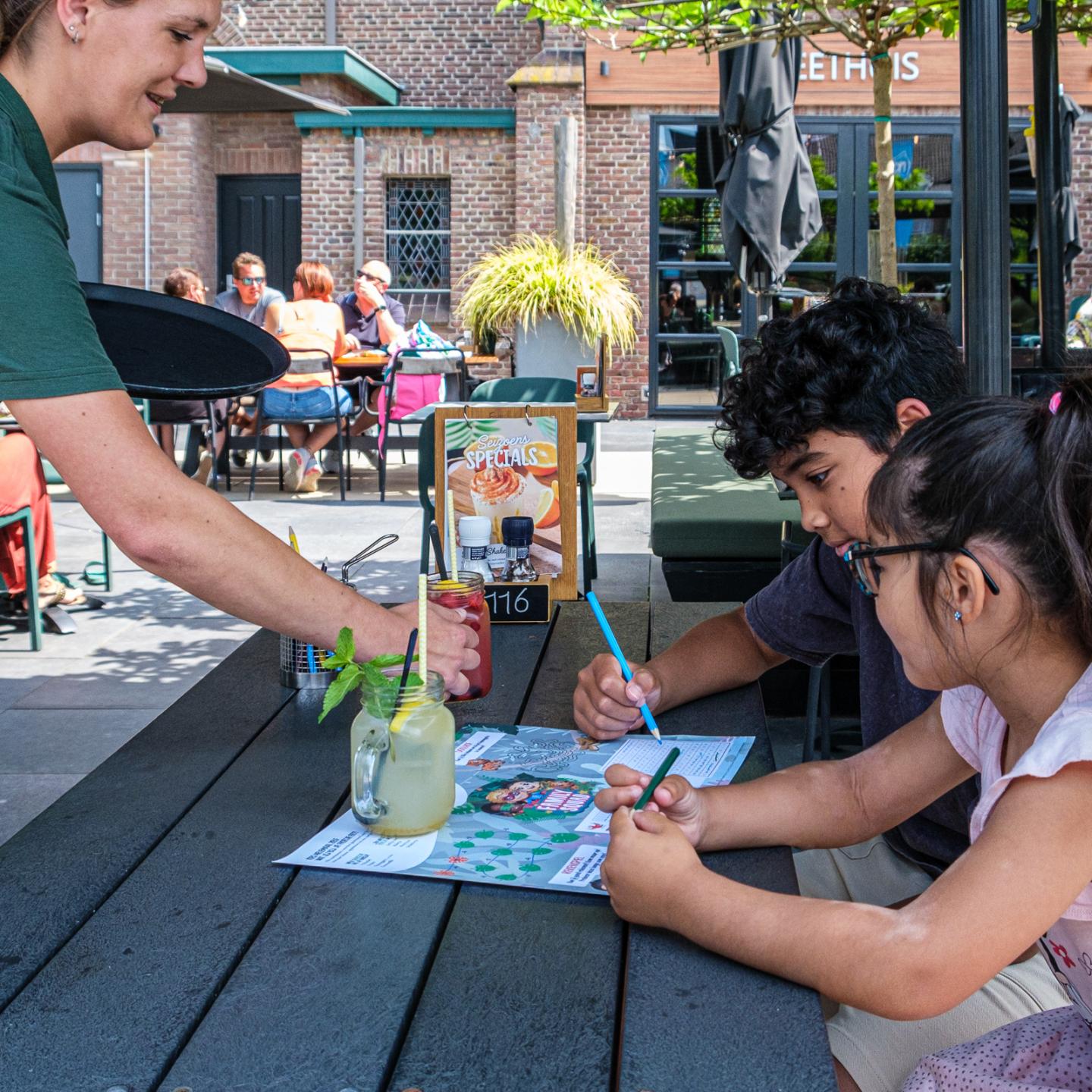 Twee kinderen zitten te kleuren aan een picknicktafel op het terras van Family Axel. Een serveerster brengt ze net een lekker drankje met een rietje