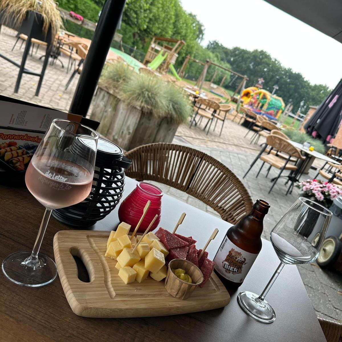 Een gedekt terrastafeltje met flesje wijn en wat hapjes met op de achtergrond de speeltuin van De Letter Asperges & Zo